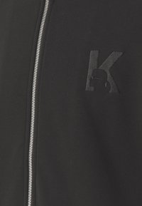 Juodas zip-up džemperis, pasižymintis tekstūruotu audiniu, sidabriniu užtrauktuku ir juodai siuvinėta raide "K" viršutiniame kairiajame krūtinės kampe.
