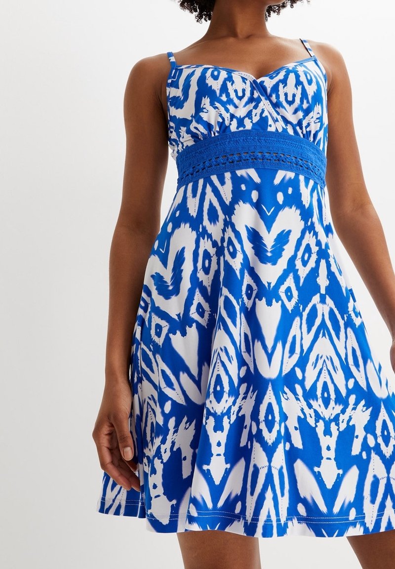 Femme portant une robe d'été sans manches à motif bleu et blanc avec une ceinture au crochet, debout devant un fond uni.