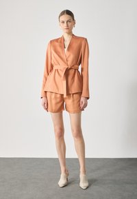 Light orange satin blazer with a tie waist, paired with matching tailored shorts. Model wears woven beige heels. Smooth texture, structured fit.