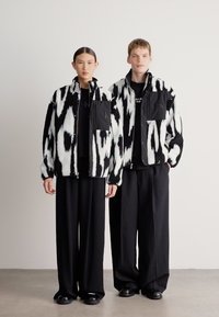 Fekete-fehér mintás dzsekik elülső zsebekkel, fekete felsők fölött viselve, széles fekete nadrággal és fekete cipővel párosítva. Simított, texturált anyag.