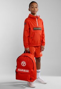 Chaqueta cortaviento de nylon roja con logo, pantalones cortos a juego, zapatillas blancas y una mochila roja con logo y detalles de cremallera.