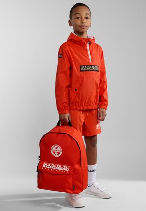 Rode nylon windjack met logo, bijpassende shorts, witte sneakers en een rode rugzak met logo en ritsdetails.