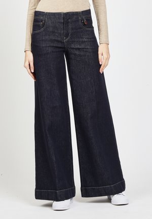 Frau trägt dunkelblaue Jeans mit weitem Bein und weiße Turnschuhe, steht vor einem schlichten Hintergrund.