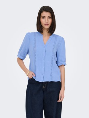 Ung kvinde iført en lyseblå knapbluse med blondefelter og mørke bukser med brede ben, poserer med den ene hånd i lommen.