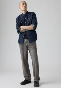 Traper košulja tamnoplave boje s kopčama na pritisak i zapadnjačkim krojem yokea. Sive traperice širokog kroja uparene s crnim cipelama. Jednostavna pozadina.