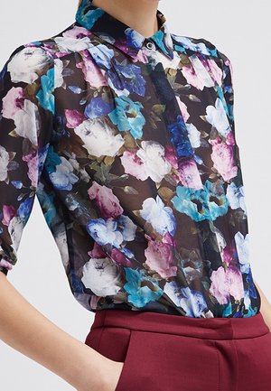 Mujer con blusa negra translúcida con estampado floral en azul, blanco y rosa, mangas abullonadas y metida dentro de pantalones de talle alto color burdeos.
