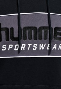 Svart sporthoodie med en grå panel som har fet "humme"-branding och texten "SPORTSWEAR", accentuerad med vita kanter och dragsnören.