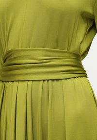 Φόρεμα σε ανοιχτό πράσινο χρώμα με πιέτες και φαρδιά ζώνη με σούρες που σφίγγει στη μέση, αναδεικνύοντας την υφή του υφάσματος και τις λεπτομέρειες των ραφών.