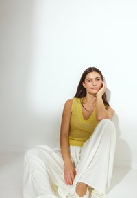 Haut sans manches en tricot jaune avec un col en V, associé à un pantalon large blanc. Textures lisses, design minimaliste sur un fond clair.