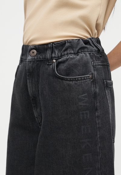 Κοντινό πλάνο μαύρου τζιν από τζιν με ελαστική μέση και αμυδρό κάθετο κείμενο στο πόδι, φορεμένο με μπεζ πουκάμισο μέσα από τη μπλούζα.