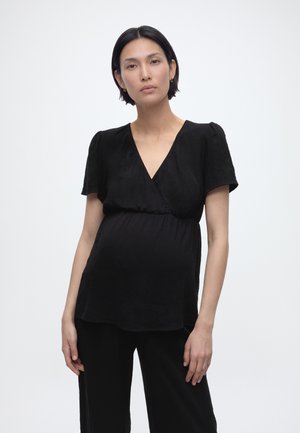 Femme enceinte aux cheveux courts et foncés, portant un haut cache-cœur noir à manches courtes et un pantalon noir, posant devant un fond blanc uni.