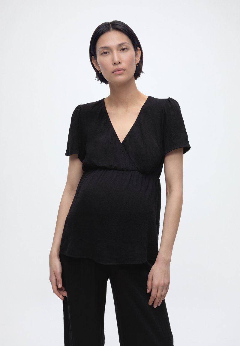 Έγκυος γυναίκα με κοντά σκουρόχρωμα μαλλιά, φοράει μαύρο κοντομάνικο τοπ τύπου wrap και μαύρο παντελόνι, στέκεται μπροστά σε απλό λευκό φόντο.