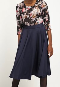 Top trasparente nero con motivo floreale e maniche a tre quarti abbinato a una gonna solid navy a forma di A, che mostra un tessuto liscio e fluente.