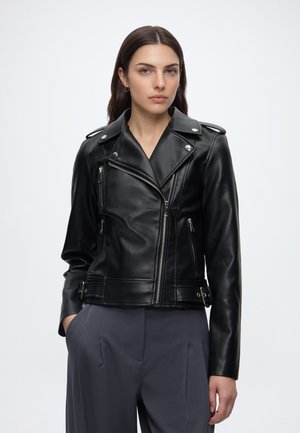 Frau mit langen dunklen Haaren, die eine schwarze Leder-Bikerjacke und dunkelgraue, hoch taillierte Hose trägt, steht vor einem schlichten weißen Hintergrund.