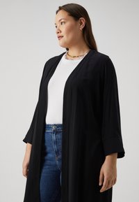 Cardigan léger de style kimono noir avec des manches trois-quarts, porté sur une chemise blanche et associé à un jean bleu. Simple, avec un devant ouvert.