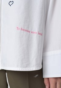 Biała koszula z długimi rękawami, z niebieskim haftem serca i różowym napisem "To forever ever land" na mankiecie. Gładka tekstura tkaniny.