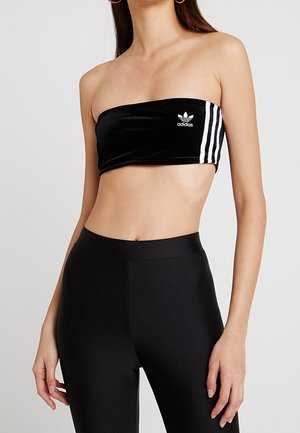 Kobieta w czarnym topie Adidas bez ramiączek z białymi paskami i dopasowanych czarnych legginsach z wysokim stanem, stoi na jednolitym tle.