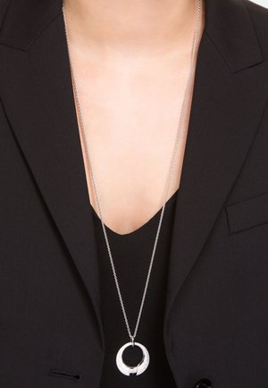 Collar de plata con un colgante circular que presenta una superficie texturizada, llevado sobre una blusa negra, complementado por un blazer oscuro.