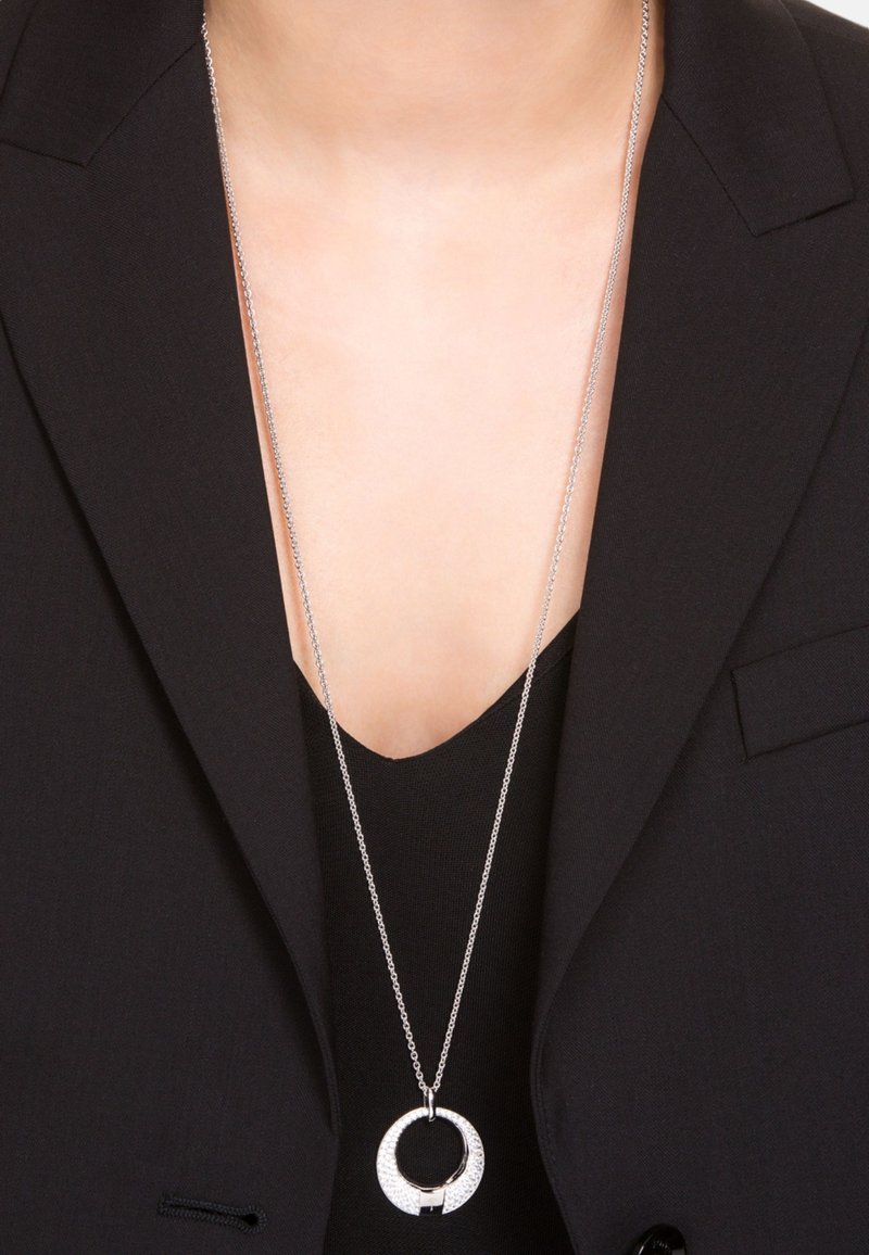 Silverhalsband med en cirkulär medaljong med en texturerad yta, bärs över en svart top, kompletterat med en mörk kavaj.