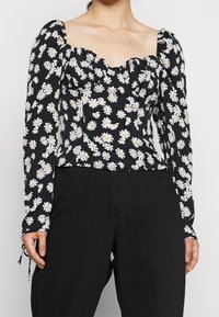 Kvinna iklädd en svart topp med vit prästkragens mönster och puffade långa ärmar, kombinerat med svarta byxor, visad från axlarna till höfterna.