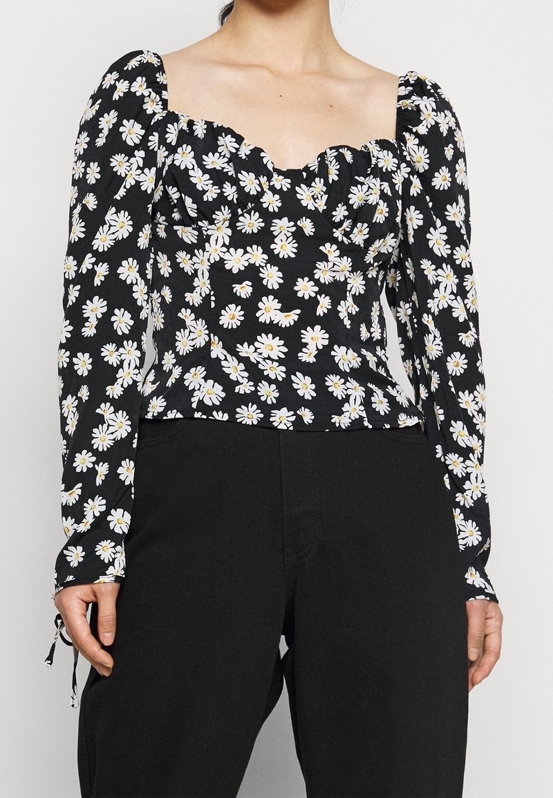 Kvinna iklädd en svart topp med vit prästkragens mönster och puffade långa ärmar, kombinerat med svarta byxor, visad från axlarna till höfterna.