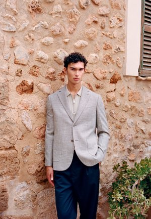 Joven con cabello rizado que lleva un blazer gris sobre una camiseta beige con cremallera y pantalones azul marino, de pie frente a una pared de piedra texturizada con vegetación.