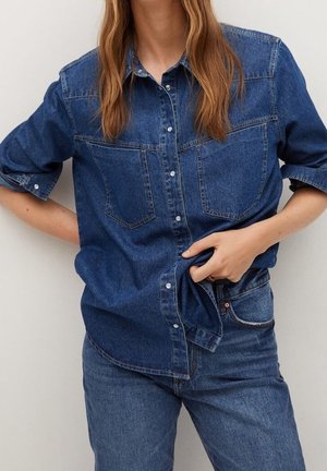 Kobieta ubrana w niebieską koszulę denim i dżinsy, poprawiająca hem koszuli prawą ręką na jednolitym jasnym tle.