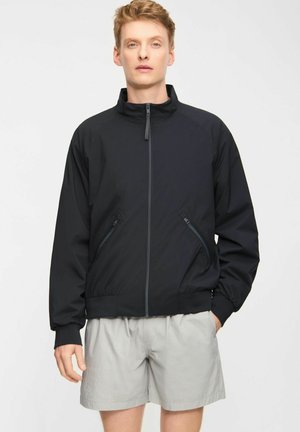 Mand iført sort lynlåstrøje og lysegrå shorts, stående foran en ensfarvet hvid baggrund.