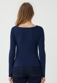 Maglia a maniche lunghe blu navy con ampia scollatura a barca, texture liscia e design aderente, abbinata a jeans scuri in denim con tasche posteriori.