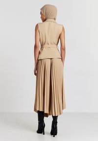 Gilet senza maniche beige con vita accentuata da una cintura e gonna plissettata, abbinato a stivali neri con tacco alto, visto da dietro.