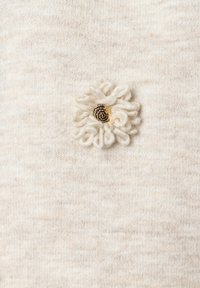 Cremfarbener Strickstoff mit einer strukturierten Oberfläche präsentiert ein dekoratives Blumenapplikation aus off-weißem Garn mit einem goldenen Mittelpunkt.