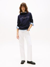 Sweatshirt marine avec le logo "TOMMY HILFIGER", manches longues, cordon argenté, superposé sur une chemise bleu clair, associé à un pantalon blanc et des mocassins noirs.