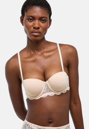 Femme aux cheveux courts portant un soutien-gorge beige rembourré avec bordure en dentelle, face à un fond clair uni.