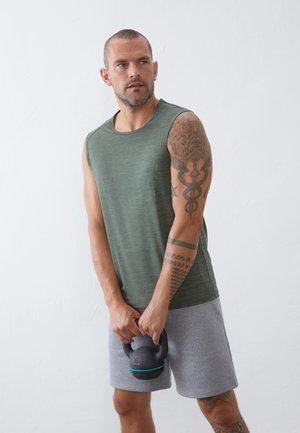 Camiseta atlética sin mangas de color verde con un patrón texturizado, pantalones cortos grises. La persona sostiene una kettlebell negra con un acento en color aguamarina.