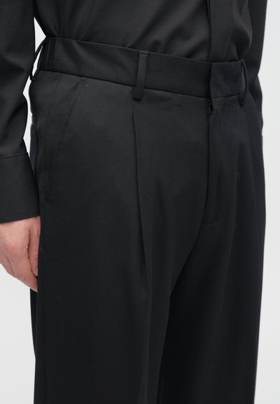 Μαύρο παντελόνι σε στενή γραμμή με λεία υφή, διαθέτει πτυχώσεις στο πλάι και ζώνη με υφή. Χωρίς ορατό μεταλλικό εξάρτημα ή σχέδια.