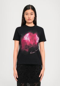 Black cotton t-shirt featuring a pink graphic design on the front, with text at the bottom. Model wears a black lace skirt.