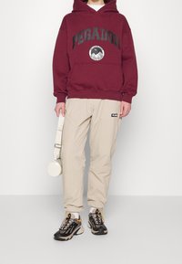 Osoba oblečená v burgundské mikině s nápisem "PEGADOR", béžových cargo kalhotách, černých a žlutých teniskách, držící malou kulatou bílou tašku za popruh.