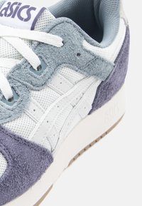 Fehér sneaker szürke és lila velúr részletekkel. Hálós anyag, kerek fűzők és párnázott bokarész. Fel van tüntetve a márkajelzés a nyelvén, valamint texturált talpa van.