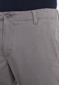 Primo piano di pantaloni grigi con passanti per cintura, chiusura a bottone e tasche laterali indossati con una maglietta scura.