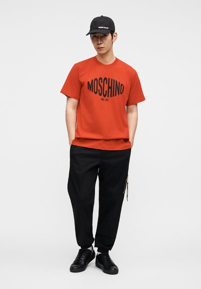 Κόκκινο βαμβακερό t-shirt με έντονη μαύρη επιγραφή "MOSCHINO", σε συνδυασμό με άνετο μαύρο παντελόνι και μαύρα αθλητικά παπούτσια. Το καπέλο προσθέτει μια σπορ λεπτομέρεια.