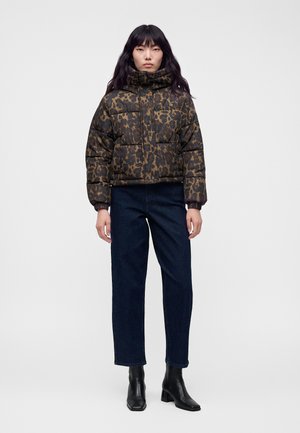 Donna che indossa una giacca corta imbottita con stampa leopardata, jeans blu scuro e stivaletti neri, in piedi davanti a uno sfondo semplice.