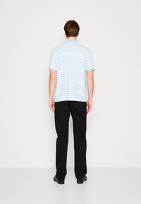 Man som står vänd bort, iförd en ljusblå och vit randig kortärmad polo, svarta jeans och svarta skor mot en enfärgad bakgrund.