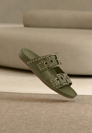 Zelený semišový šľapák s dvoma striebornými prackami, cvočkami a plochou podrážkou. Pozadie zobrazuje mäkké, neutrálne tóny.