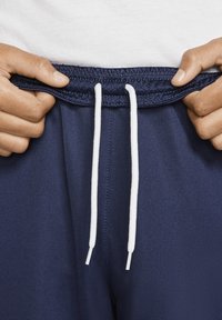 Manos ajustando la cinturilla de unos pantalones deportivos azul marino con cordones blancos, contra un fondo de camisa blanca.