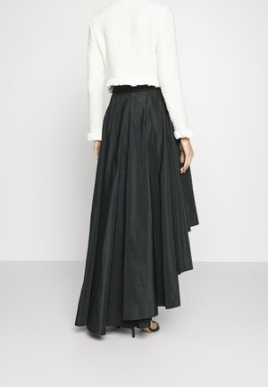 Kobieta w długiej czarnej asymetrycznej spódnicy i białej teksturowanej bluzce z długim rękawem, widziana od tyłu, stojąca na jednolitej podłodze.