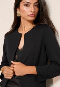 Czarna marynarka z okrągłym dekoltem, dopasowany krój, długie rękawy i gładka faktura, noszona na koronkowej bluzce. Złote bransoletki podkreślają nadgarstki.