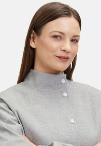 Grijze, gestructureerde jas met een hoge kraag, voorzien van twee ronde knopen en een gestructureerd ontwerp. De stof lijkt zacht en lichtgewicht te zijn.