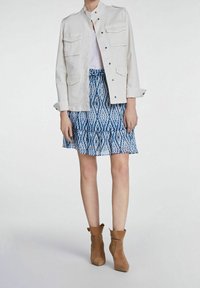 Veste en denim blanc avec poches à rabats, superposée à une robe bleue à motifs avec des volants, assortie à des bottines brunes.