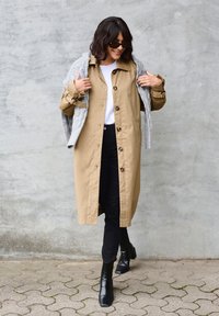 Béžový trenčkot s gombíkmi, skombinovaný so šedým pleteným šálom. Čierne nohavice a čižmy na vysokom opätku dotvárajú outfit.