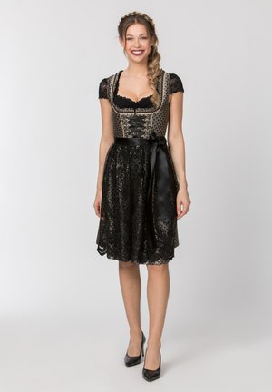 Junge Frau lächelt, trägt ein schwarzes und goldenes traditionelles Dirndl mit Spitzenärmeln und einer Satinschleife in der Taille, steht vor einem einfarbigen Hintergrund.
